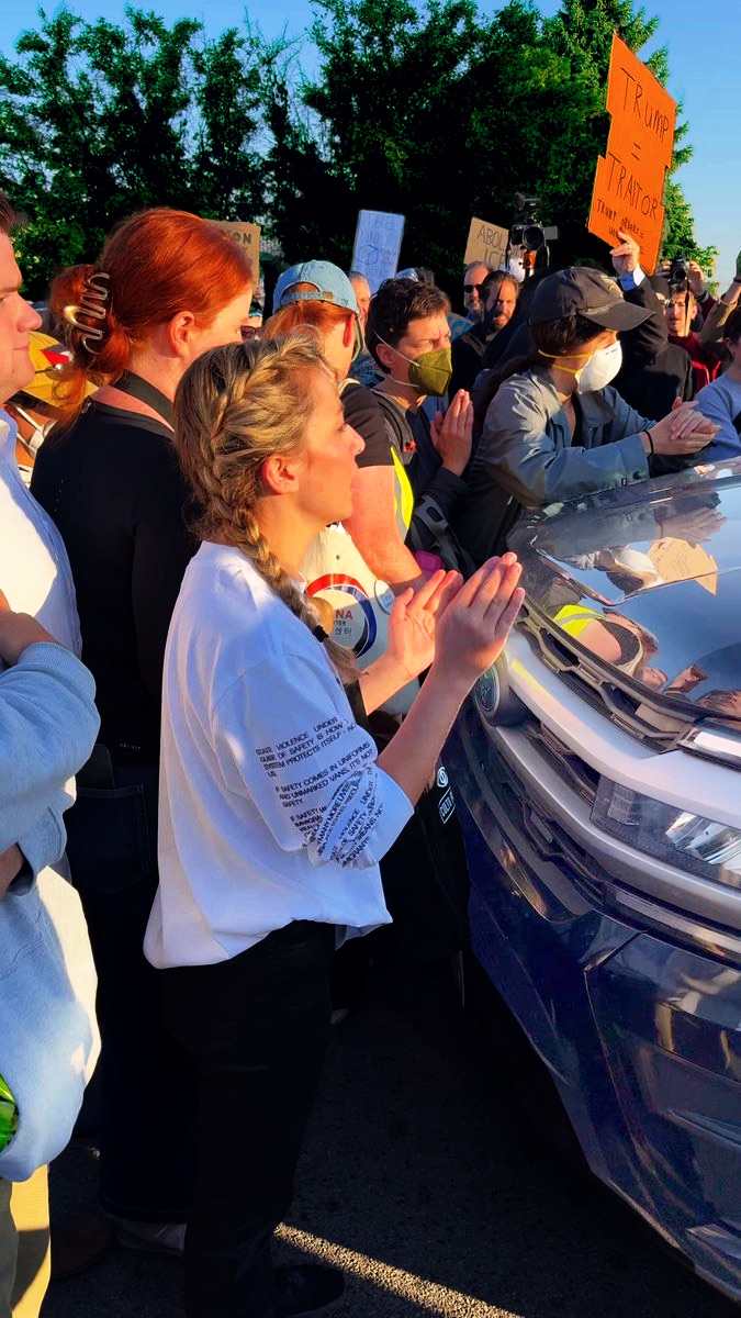 A crowd of protesters presses against the hood of a black SUV in low evening light; a woman with braided hair at the front of the hood holds her hands up against it.